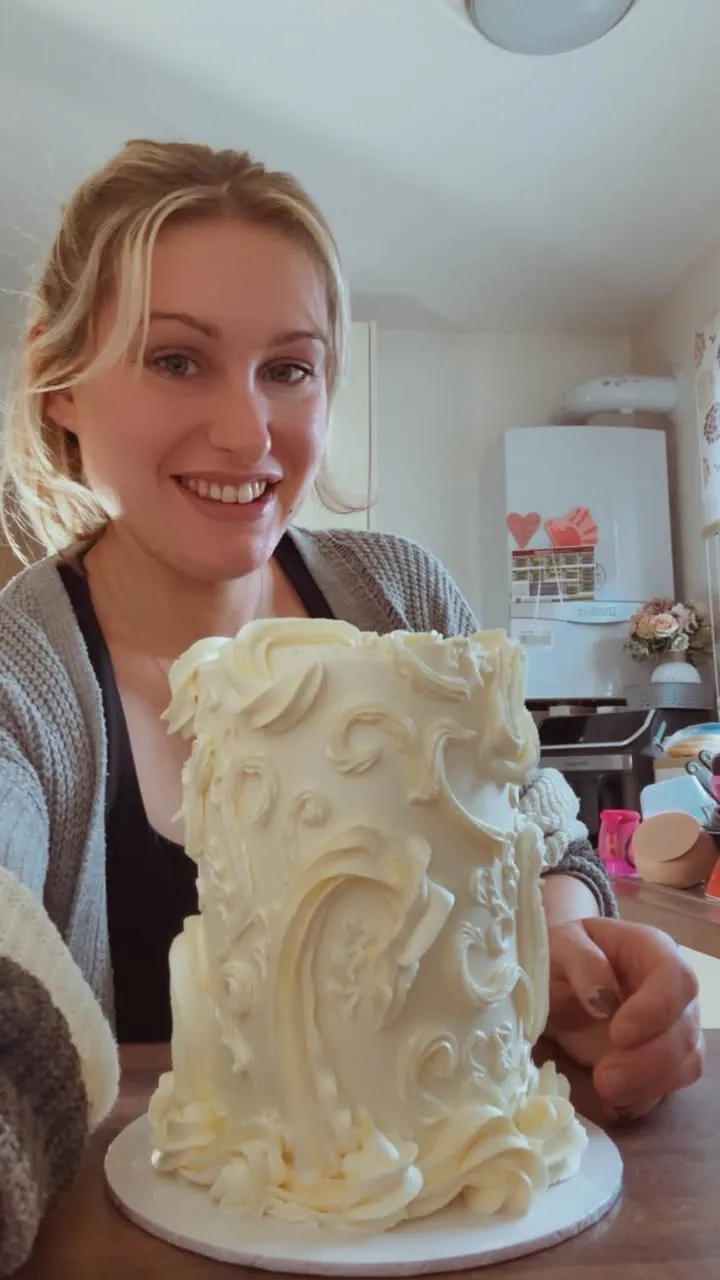 Dana, the baker, smiling behind a tall cake covered in piped white buttercream